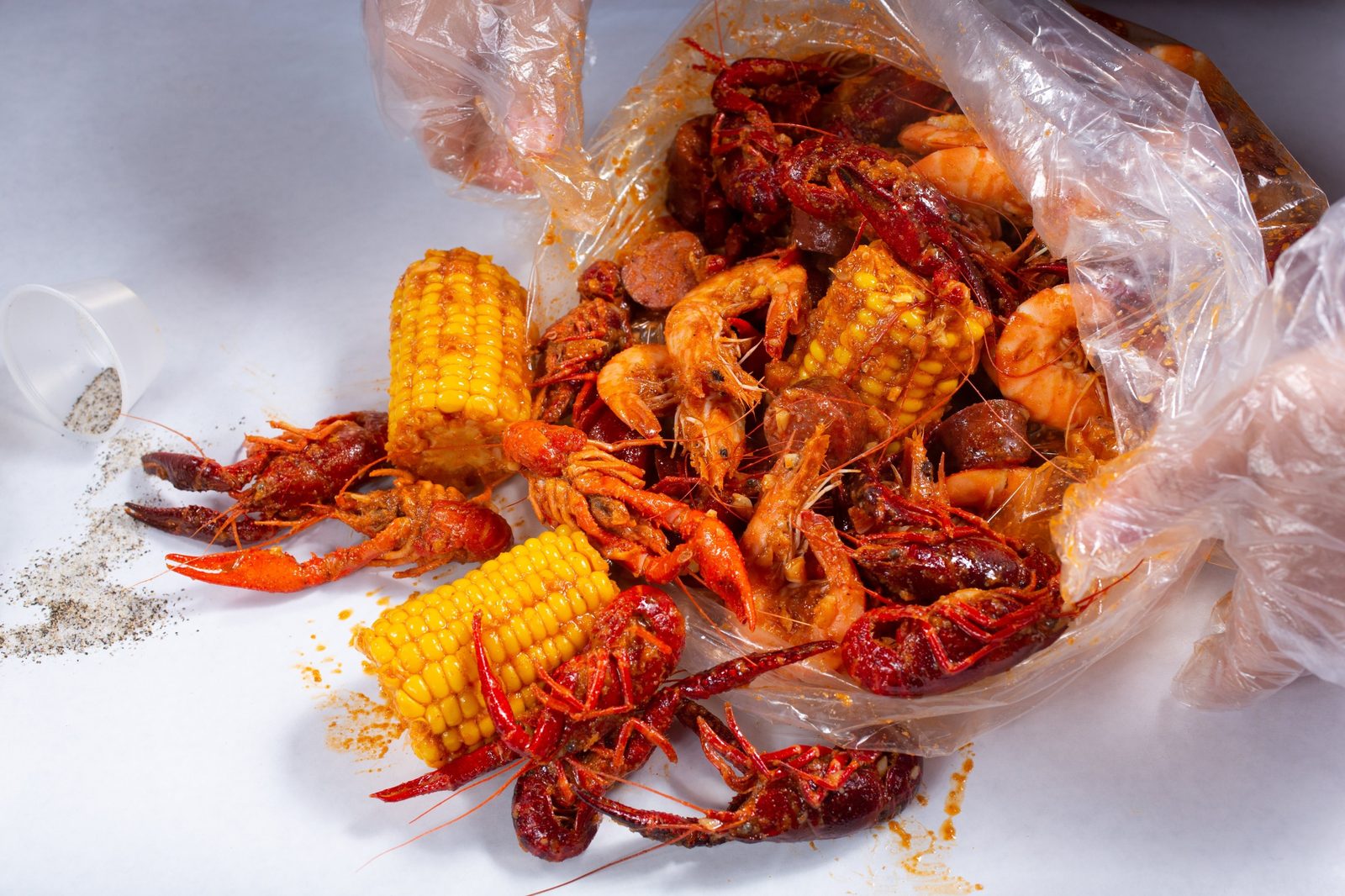 Gloved hands pouring a saucy shrimp, crawfish and corn boil from a clear bag